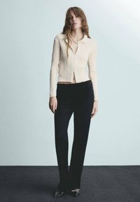 Cardigan a coste crema con maniche lunghe e colletto, abbinato a pantaloni neri su misura e décolleté neri a punta, su uno sfondo chiaro.