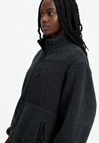 Giacca in pile grigio scuro con pannello frontale in nylon nero liscio, tasche con zip, collo alto e tessuto strutturato per il calore.