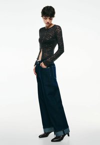 Haut long à manches en dentelle noire avec un design �à boutons à l'avant, associé à un jean en denim foncé à taille haute et jambes larges, avec des poignets retroussés et une coupe classique.