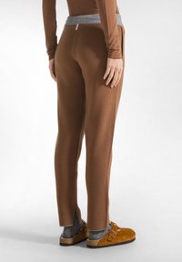 Bruine fleece broeken met een grijze tailleband, met een aansluitende pasvorm en zijsplitten bij de enkels. Gedragen met beige instappers en grijze sokken.