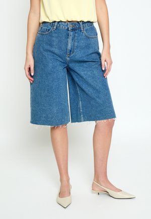 Osoba nosząca niebieskie jeansowe szorty o wysokim stanie sięgające do kolan z surowymi wykończeniami, zestawione z beżowymi sandałkami na obcasie typu slingback i włożonym żółtym topem.
