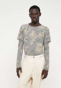 Tricou gri camuflaj cu mânecă lungă, decolteu rotund și croială lejeră, asortat cu blugi largi de culoare deschisă, cu cusături decorative.