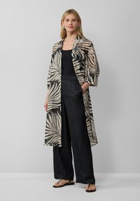 Leichter, transparenter langer Mantel mit einem schwarz-beigen Blättermuster über einem schwarzen Top, kombiniert mit schwarzen weiten Hosen und Flip-Flops.