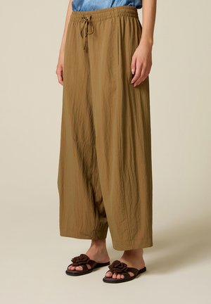 Mujer con pantalones marrones sueltos de cintura alta con cinturilla elástica y sandalias marrones adornadas con decoraciones de flores.