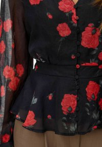 Mustapohjainen kukkatoppi, jossa on punertavia unikkokuvioita, pitkät läpikuultavat hihat, peplum-vyötärö sekä kuusi nappisuljinta edessä.