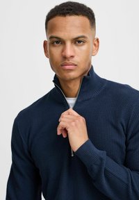 Mornarska zip-up majica od teksturiranog materijala, s rebrastim ovratnikom i manžetama. Model drži patentni zatvarač ispred neutralne pozadine.