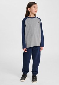 Graues Langarmshirt mit dunkelblauen Raglanärmeln und einem kleinen Logo auf der Brust, kombiniert mit navyfarbigen Sweatpants und schwarzen Sneakers.