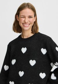 Schwarzer Pullover aus strukturiertem Stoff mit weißen Herzmustern. Klassischer Rundhalsausschnitt und lockere Passform.