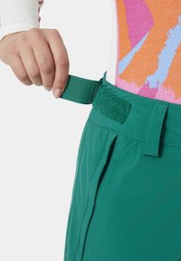 Grüne Outdoor-Hosen mit einem Klettverschluss und Reißverschlusstaschen. Der Stoff ist leicht und hat eine glatte Textur. Akzentuiert durch ein farbenfrohes gemustertes Oberteil.