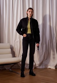 Black bomber jacket with a zip front, worn over a light green top, paired with high-waisted dark jeans and black ankle boots.