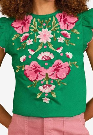 Groene blouse met grote roze bloemrijke borduursels en gerimpelde korte mouwen, gedragen met roze broek, zichtbaar van schouders tot taille.