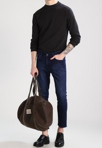 Dark brown canvas duffel bag with two sturdy handles, circular shape, and a label on the front; worn with dark jeans and a black long-sleeve top.