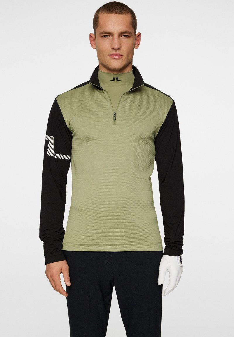 Tight passform långärmad tröja med halv dragkedja. Kroppen i olivgrön, svarta ärmar, texturerad tyg och vitrandig logotyp på vänstra ärmen.