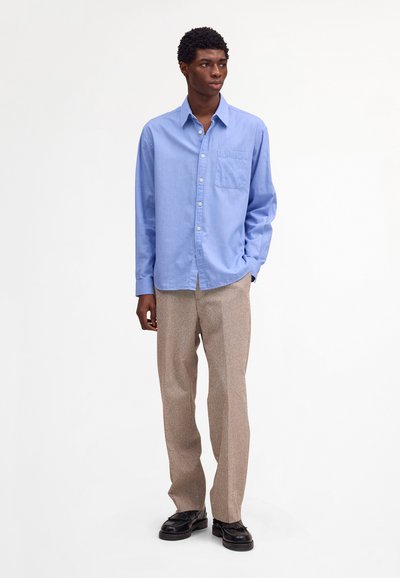 Uomo in piedi con mano nella tasca, indossa una camicia azzurro chiaro con bottoni, pantaloni beige e mocassini neri su sfondo bianco uniforme.