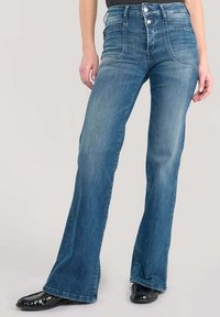 Jeans en denim bleu évasés avec une taille haute, présentant des poches avant, une texture lisse et des boutons-pression en argent. Chaussures noires visibles au bas.
