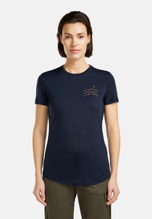 Frau trägt ein marineblaues T-Shirt mit kurzen Ärmeln und einem kleinen orangefarbenen Design eines Berges und einer Sonne auf der linken Brust, kombiniert mit olivgrünen Hosen.
