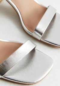 Metalliska silverklackar med en bred rem över toppen, fyrkantig tåform och slät textur på en tanfärgad innersula.