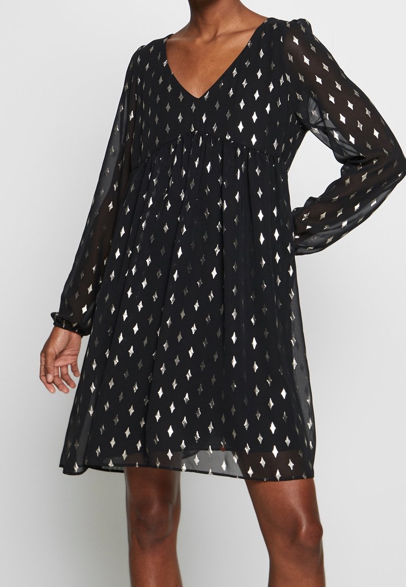 Robe noire à col en V avec de longues manches transparentes et des motifs métalliques argentés en forme de losange, portée par une personne debout, une main sur la hanche.