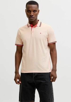 Homme portant un polo rose clair avec un col rayé rouge et noir et un short noir, debout devant un fond uni clair.