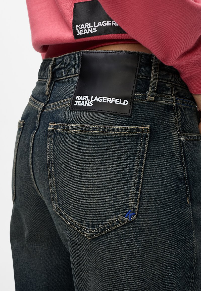 Dunkle Denim-Jeans verfügen über ein schwarzes Lederetikett mit der Aufschrift "KARL LAGERFELD JEANS" in weißem Text, feinen Nähten und einem kleinen blauen Akzent.