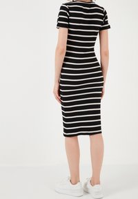 Donna in piedi di spalle che indossa un vestito a righe orizzontali bianche e nere di lunghezza ginocchio e sneakers bianche su uno sfondo bianco semplice.