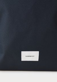 Granatowa torba z płótna z białą prostokątną metką z logo "SANDQVIST", prezentująca teksturowaną powierzchnię tkaniny. Prosty, funkcjonalny design.