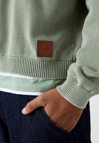 Zielony sweter z dzianiny z prążkowanymi mankietami i szwem, z brązową skórzaną naszywką z napisem "GUESS JEANS 1981." W zestawie z ciemnymi jeansami.