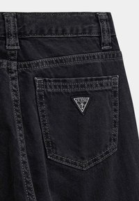 Jeans in denim nero con una tasca posteriore che presenta un'etichetta a forma triangolare con il logo "GUESS". Le cuciture scure contrastano con la trama del tessuto.
