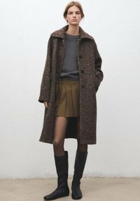 Manteau long texturé marron avec grand col et boutons sur un pull gris, associé à une jupe plissée marron et des bottes noires montantes jusqu'aux genoux.