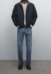 Navy bomber jacket with a gray collar, beige knitted sweater, light blue jeans, and black shoes with brown laces. Simple and clean design.