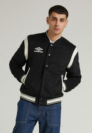 Veste varsity noire avec un logo blanc, des manches crème contrastantes et des poignets côtelés rayés, dotée d'une texture douce et de boutons-pression.