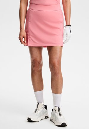 Kvinde iført en pink golfskørt, hvid golfhandske på venstre hånd, hvide sokker og hvide sneakers stående på en ensfarvet baggrund.