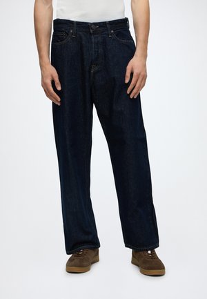 Homme portant un jean droit bleu foncé, une chemise blanche et des baskets marron, debout devant un fond uni.