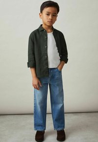 Camisa verde de botones sobre una camiseta blanca de tirantes, combinada con jeans de un azul claro y zapatos marrones oscuros. Diseño simple, corte relajado y textura media.