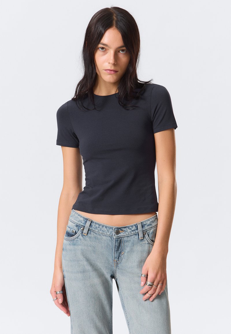 Schwarzes, kurzes T-Shirt aus einem soliden, glatten Stoff. Kurze Ärmel und ein runder Ausschnitt. Kombiniert mit hellblauen, leicht verwaschenen Jeans.