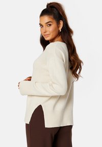 Maglione beige chiaro con scollatura rotonda, polsini a coste e spacchi laterali. Tessuto morbido, vestibilità rilassata, indossato sopra pantaloni marroni scuri.