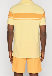 Polo-shirt in lichtgeel met oranje horizontale strepen. Katoenen stof, korte mouwen, gecombineerd met bijpassende oranje shorts.