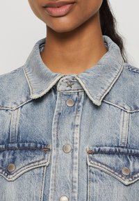 Απαλή μπλε τζιν ζακέτα με κλασικό γιακά, διαθέτοντας ασημί κουμπιά με σ snap και δύο τσέπες στο στήθος. Με φθαρμένες άκρες και ορατή ραφή.