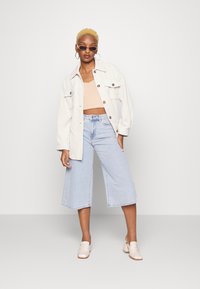 Casaco de corduroy bege claro, top crop pêssego, culottes de denim largas azuis e sapatos slip-on cremes. Os acessórios incluem óculos de sol ovoides e brincos em argola.