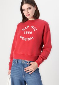 Crvena dukserica s bijelim natpisom "GAP NYC 1969 ORIGINAL", rebrasti ovratnik i manžeti, u kombinaciji s plavim trapericama visokog struka. Jednostavan, ležeran dizajn.