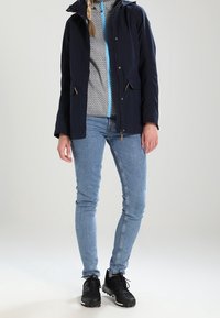 Námořnická voděodolná bunda s kapucí, dvěma předními kapsami, v kombinaci se světle modrými džíny a černými teniskami, s vzorovanou vnitřní vrstvou.