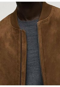 Homme portant une veste bomber en daim marron par-dessus un pull en maille gris, fermeture éclair partiellement ouverte, visible du cou à la poitrine.