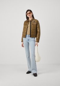 Giacca trapuntata verde oliva con due tasche sul petto, abbinata a una maglietta beige e jeans a campana azzurri chiari, con una borsa bianca. Ballerine marroni.