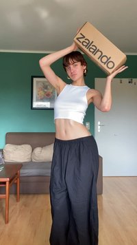 Καφέ χαρτοκιβώτιο με το λογότυπο "zalando" τυπωμένο σε μαύρο χρώμα, κρατημένο πάνω από το κεφάλι από ένα άτομο που φοράει άσπρο crop top και μαύρο φαρδύ παντελόνι.