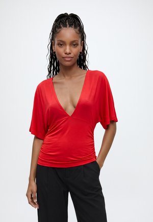 Femme aux cheveux tressés portant des boucles d'oreilles en or en forme d'anneau, un haut rouge à décolleté en V profond et un pantalon noir, debout avec une main dans la poche.