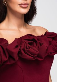 Bordowa sukienka z odkrytymi ramionami, zdobiona dużymi, teksturowanymi różami z tkaniny wzdłuż dekoltu oraz gładkim gorsetem.