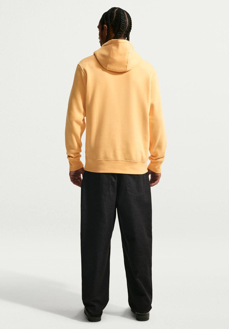 Mann mit geflochtenem Haar, der ein helloranges Kapuzensweatshirt und lockere schwarze Hosen trägt, steht mit dem Rücken zu vor einem einfarbigen Hintergrund.