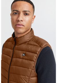 Braune gepolsterte Weste aus einem glänzenden Material, mit hohem Kragen und schwarzem Reißverschluss, die über einem dunkelblauen strukturierten Pullover getragen wird.