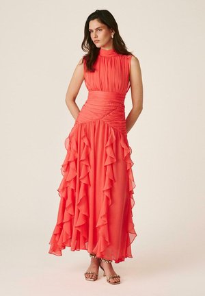 Mujer con vestido coral sin mangas con falda con volantes y cintura fruncida, de pie con las manos detrás de la espalda sobre fondo beige.