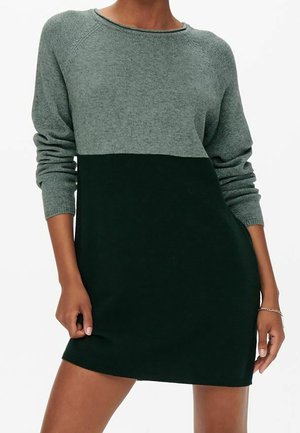 Femme portant une robe en maille bicolore à manches longues avec un haut gris et un bas noir, debout avec une main près de l’ourlet.
